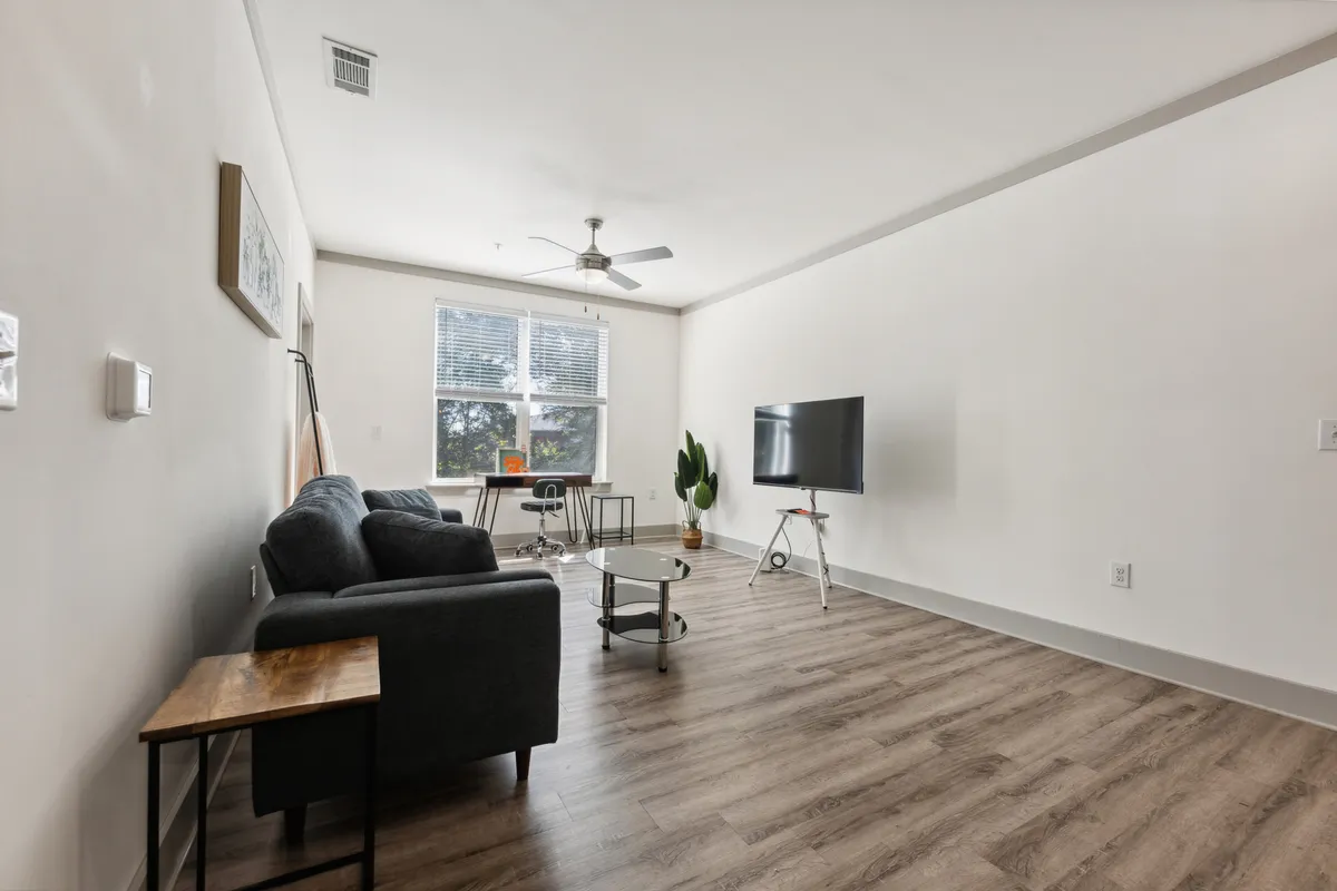 This image presents the central living area, featuring a stylish sofa, coffee table, and a smart TV. Large windows invite natural light, and the arrangement includes a workspace, hinting at both relaxation and productivity. The decor is contemporary and the ambiance is relaxed, aligning with the narrative of a luxe urban sanctuary.