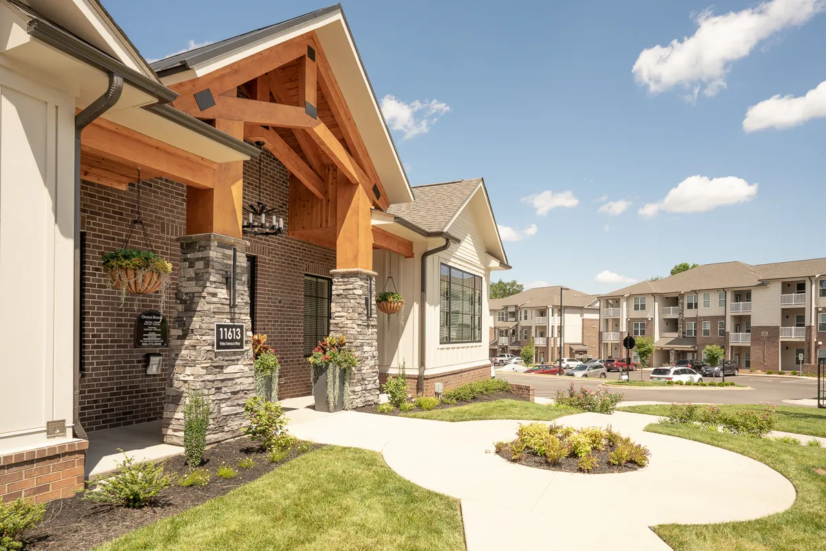 A welcoming exterior entrance featuring a distinctive wooden arch, expansive windows, colorful flower planters, and manicured landscaping. The scene evokes the first impression of arriving at Overlook 3111, seamlessly blending residential comfort with urban accessibility.