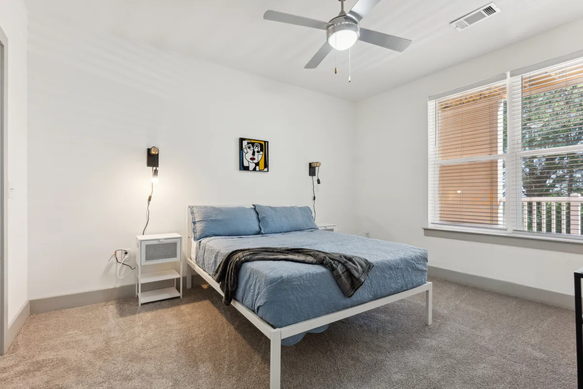 A serene bedroom featuring a plush bed dressed in crisp linens, a nightstand with a lamp, artwork on the wall, and a large window that fills the room with natural light. The ambiance is peaceful and comfortable, ideal for relaxation and restful sleep.