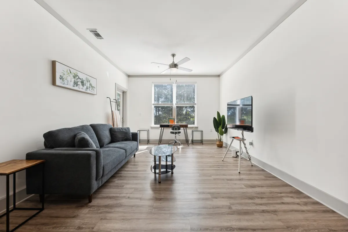 The living room here is bathed in natural light, with a comfortable sofa, coffee table, TV, and touches of greenery. The room appears inviting for reading, socializing, or simply unwinding, and the modern decor supports an atmosphere of calm and comfort.