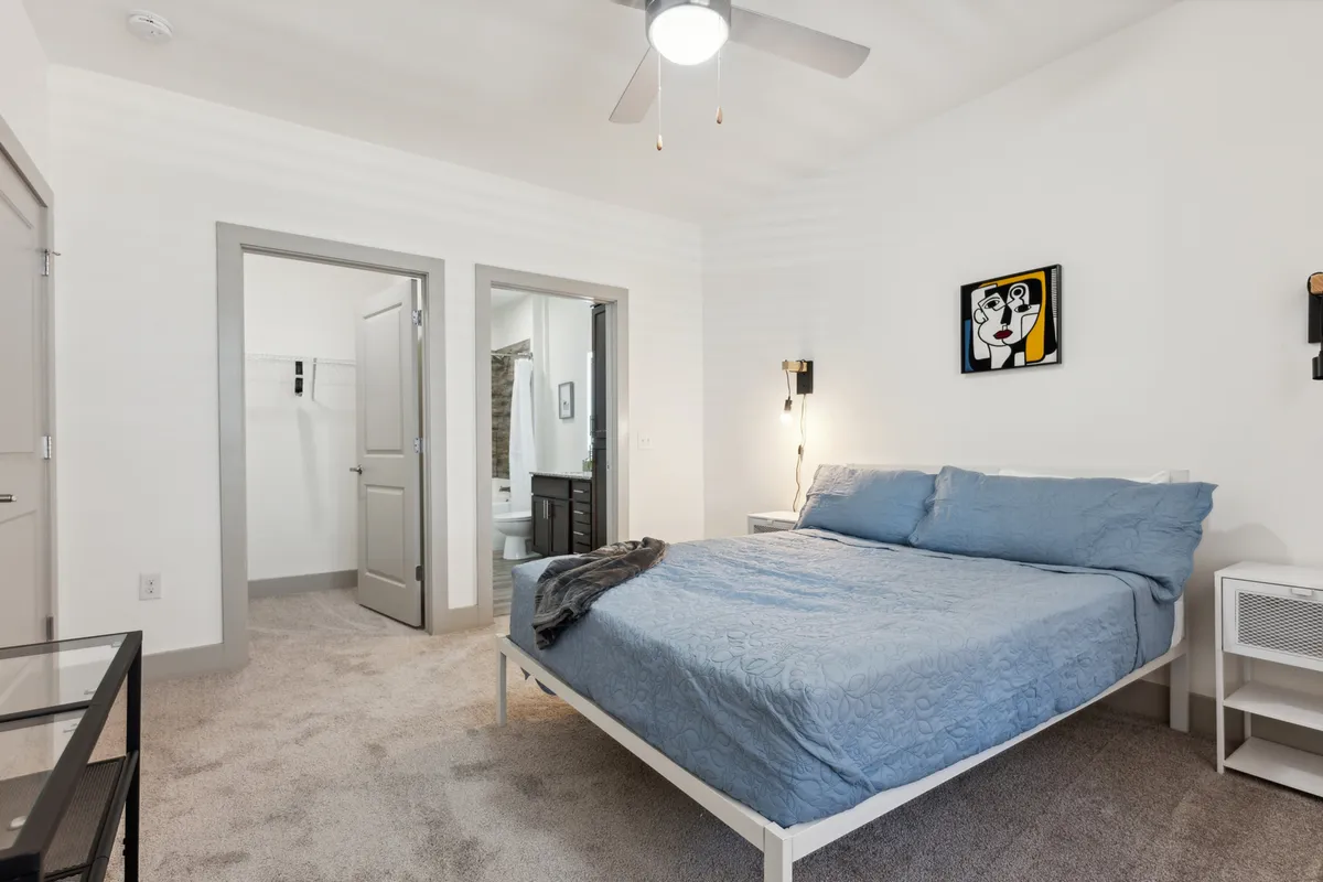 A cozy bedroom with blue bedding, modern artwork, a nightstand with a lamp, and open doors that reveal the adjacent bathroom and closet. The space feels intimate and functional, emphasizing comfort and seamless access to amenities.