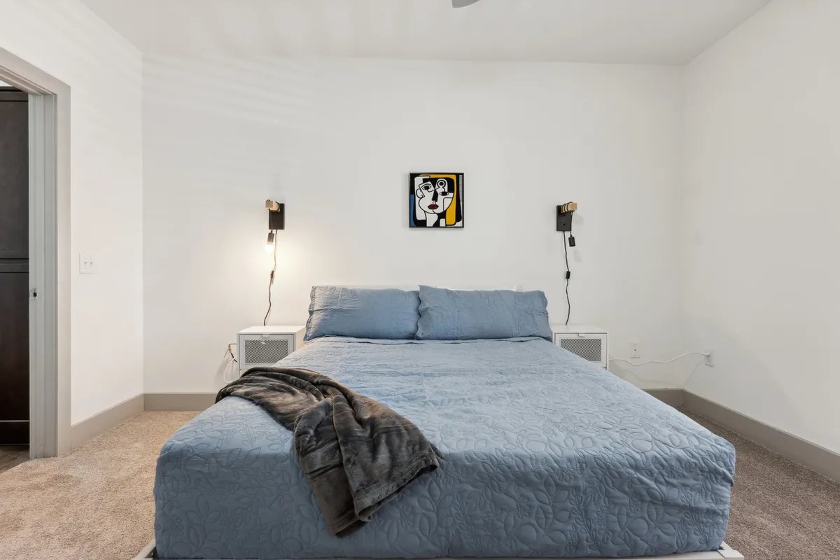 The image showcases a serene bedroom featuring a plush bed dressed in inviting blue bedding, flanked by contemporary nightstands with warm, glowing lamps. Statement art adds a touch of sophistication, while soft natural light streams in, creating a tranquil and cozy ambiance ideal for relaxation.
