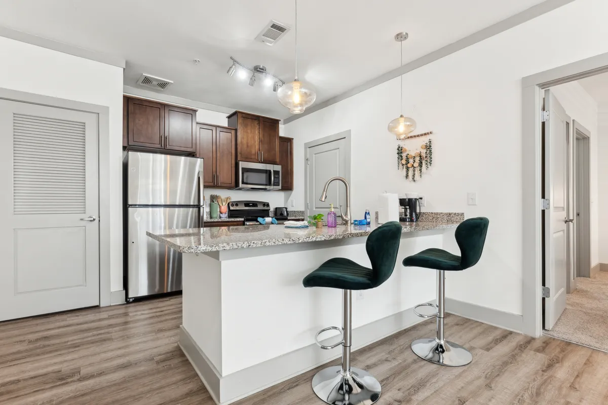 This image highlights a modern kitchen featuring a granite countertop with bar stools, sleek stainless steel appliances, and stylish cabinetry. The space is illuminated with bright, inviting lighting, creating an atmosphere perfect for both cooking and socializing.