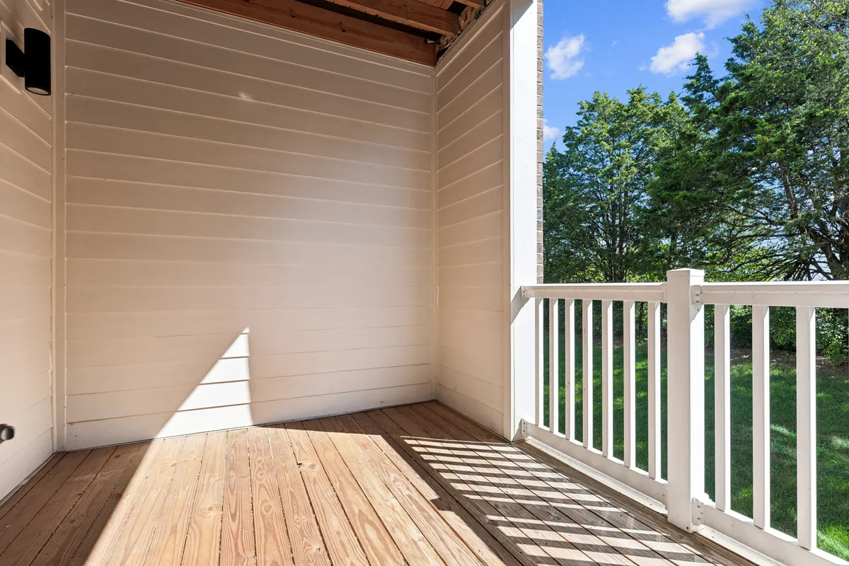 A serene exterior balcony or patio space with natural wooden floors, white railings, and abundant natural light. The setup suggests a relaxing spot to unwind, read, or enjoy a coffee while overlooking the surrounding area.